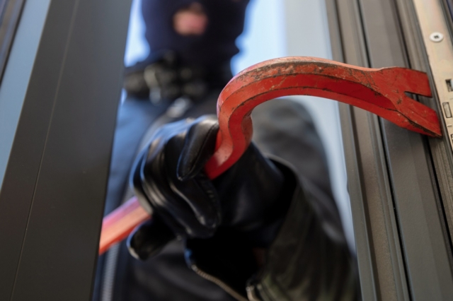 Bandit in balaclava holding a crowbar to break a glass window.
